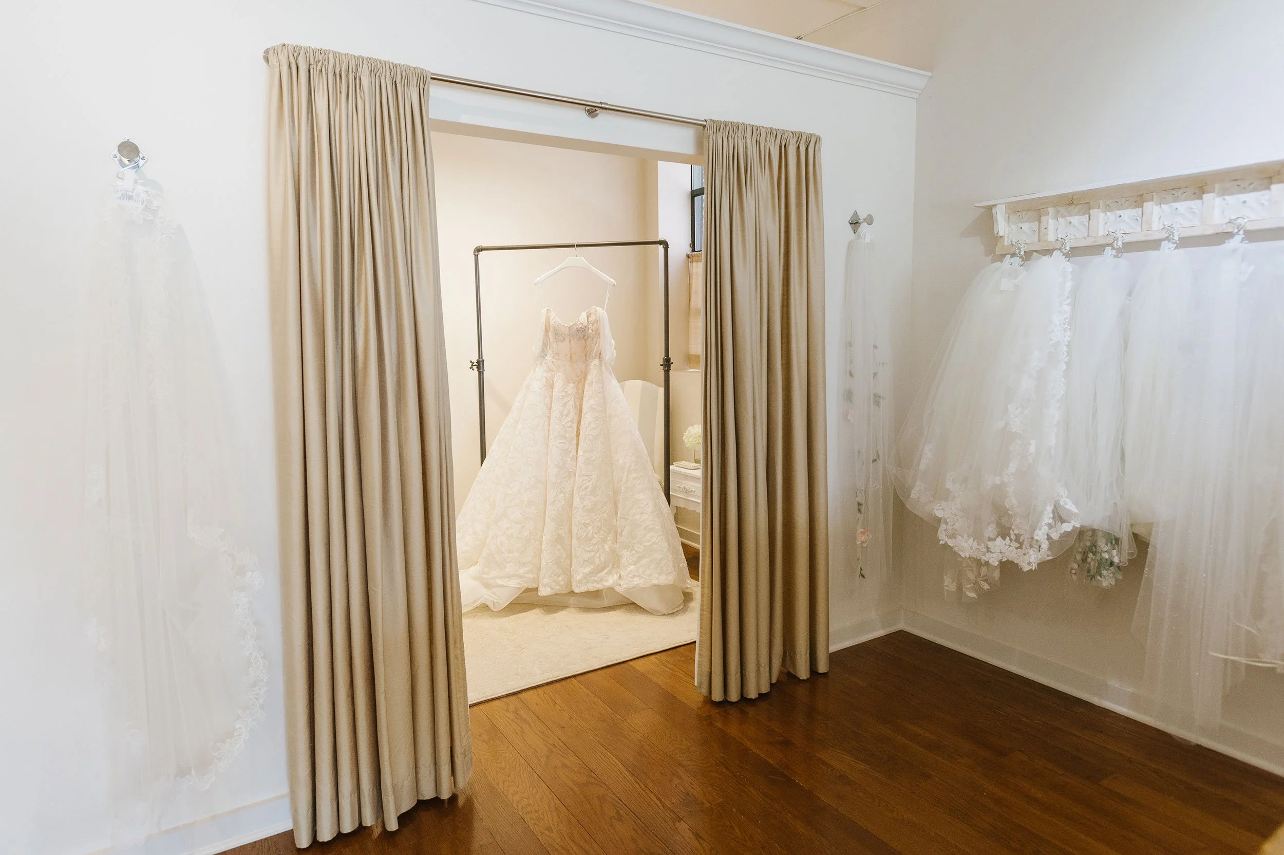 A bridal boutique fitting room with beige curtains, showcasing a wedding dress on a mannequin, and several dresses hanging on a white rack.