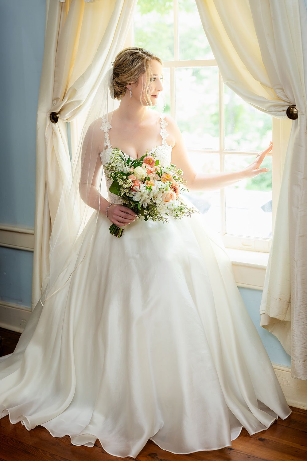 A bride in a white wedding dress holding a bouquet of flowers, standing by a window with curtains, gazing outside on a bright day.