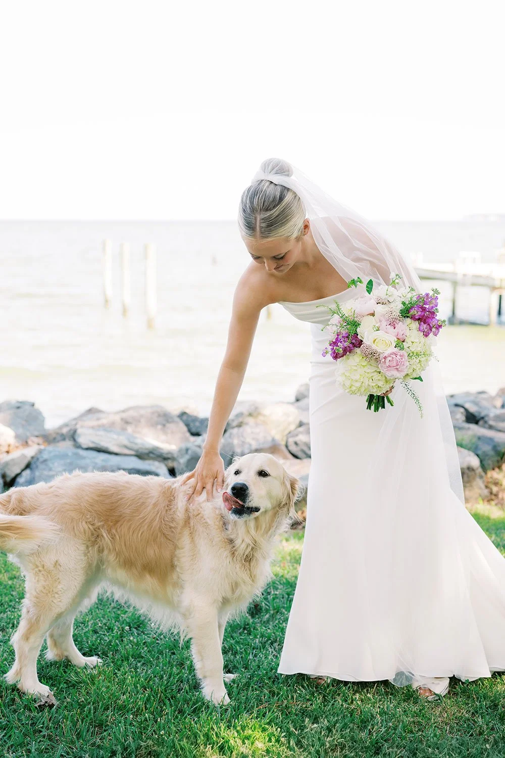 Bride in a white wedding dress and veil holding a bouquet of pink and white flowers, petting a golden retriever dog outdoors near a body of water with rocks and grass.
