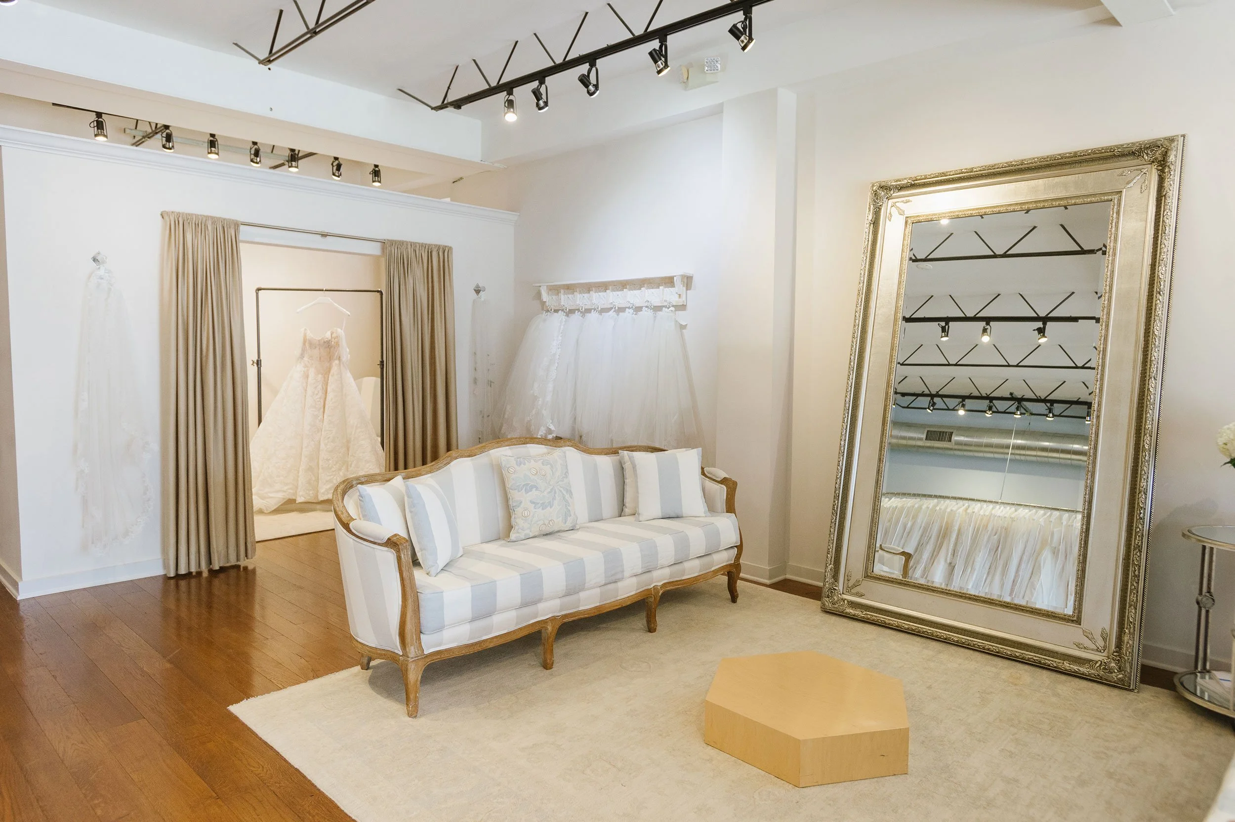 A fitting room area with a white and beige upholstered sofa, a standing mirror with an ornate gold frame, a white bridal gown on a rack, and beige curtains, in a boutique or showroom setting.