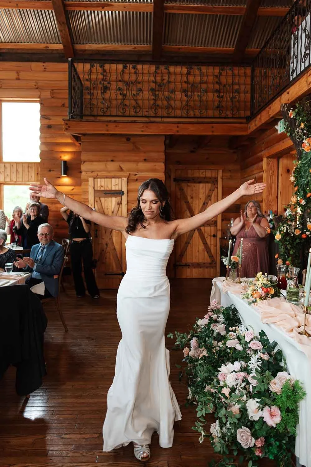 Bride in a white strapless wedding dress dancing with arms outstretched in a rustic wooden wedding reception hall decorated with flowers.