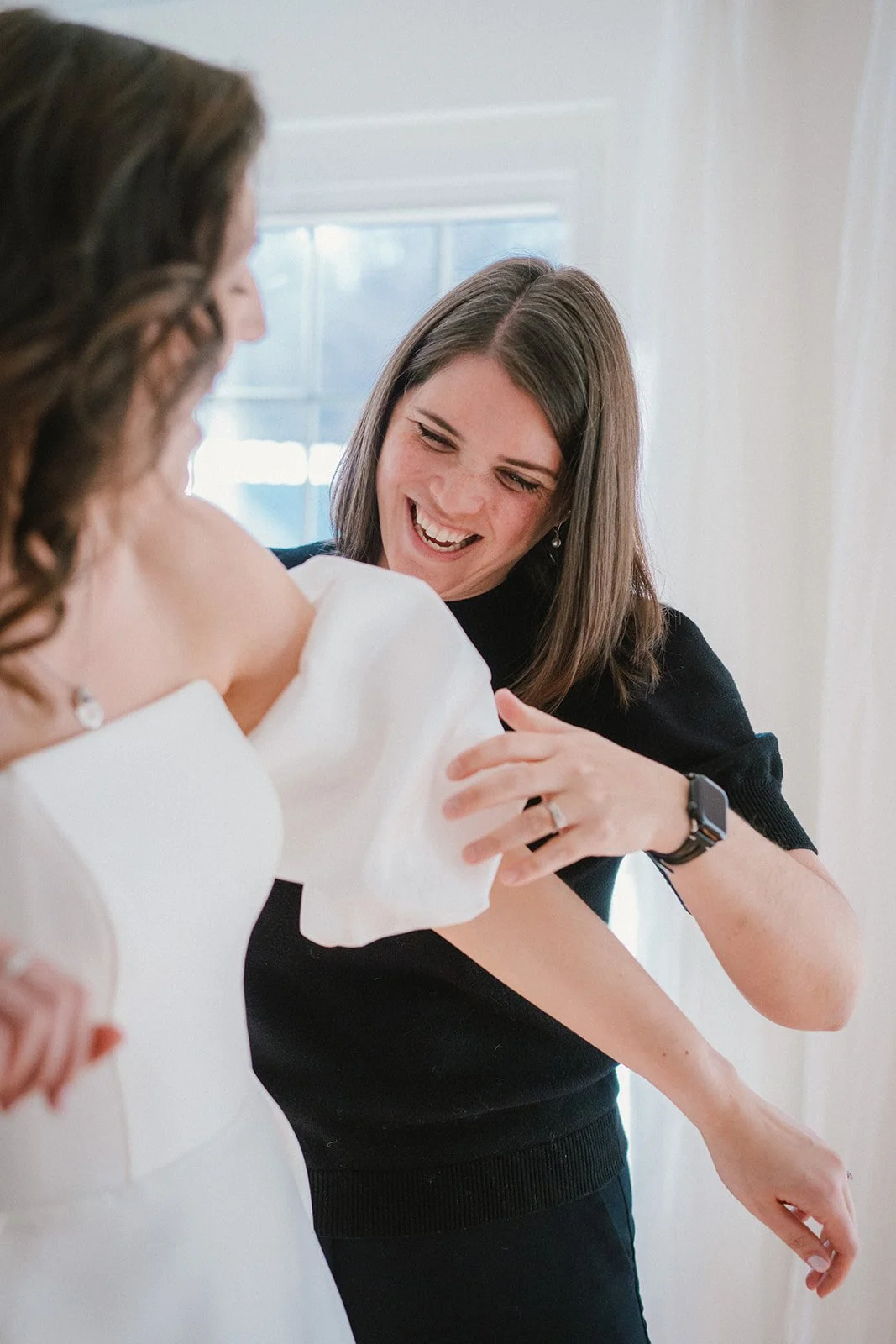 A smiling woman is helping another woman adjust a wedding dress, standing near a window with white walls.