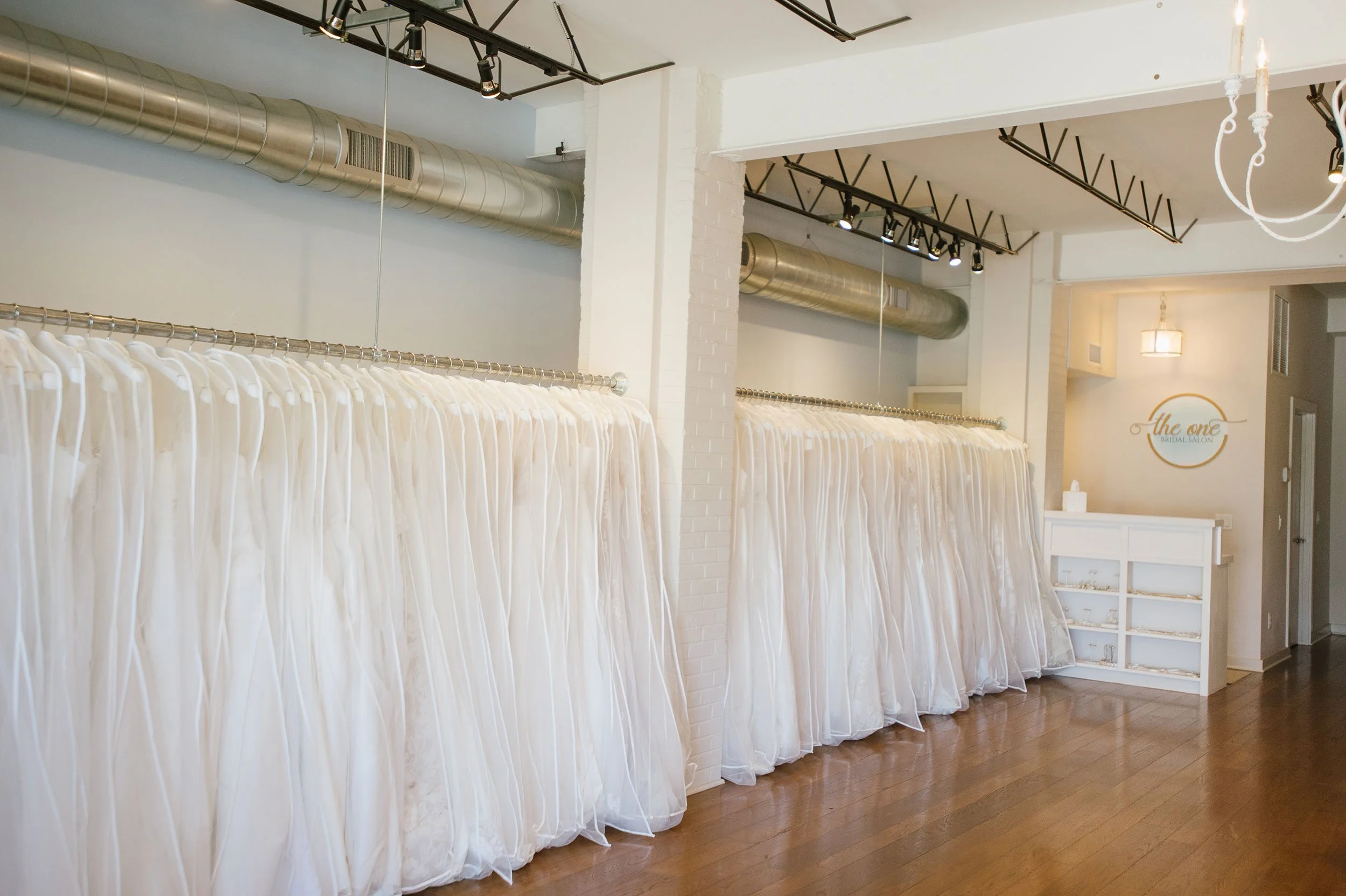 Interior of a bridal salon with white dresses on a rack, exposed ductwork on the ceiling, track lighting, a white reception desk, and a sign that reads "the one bridal salon" on the wall.