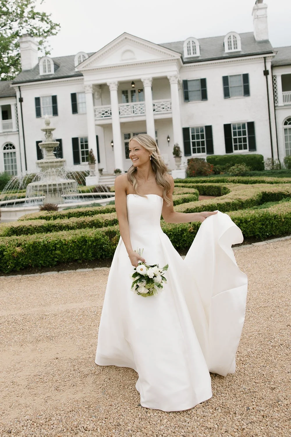 A bride in a white wedding dress holding a bouquet of flowers, standing on a gravel pathway in front of a large white mansion with black shutters, a fountain, and landscaped gardens.