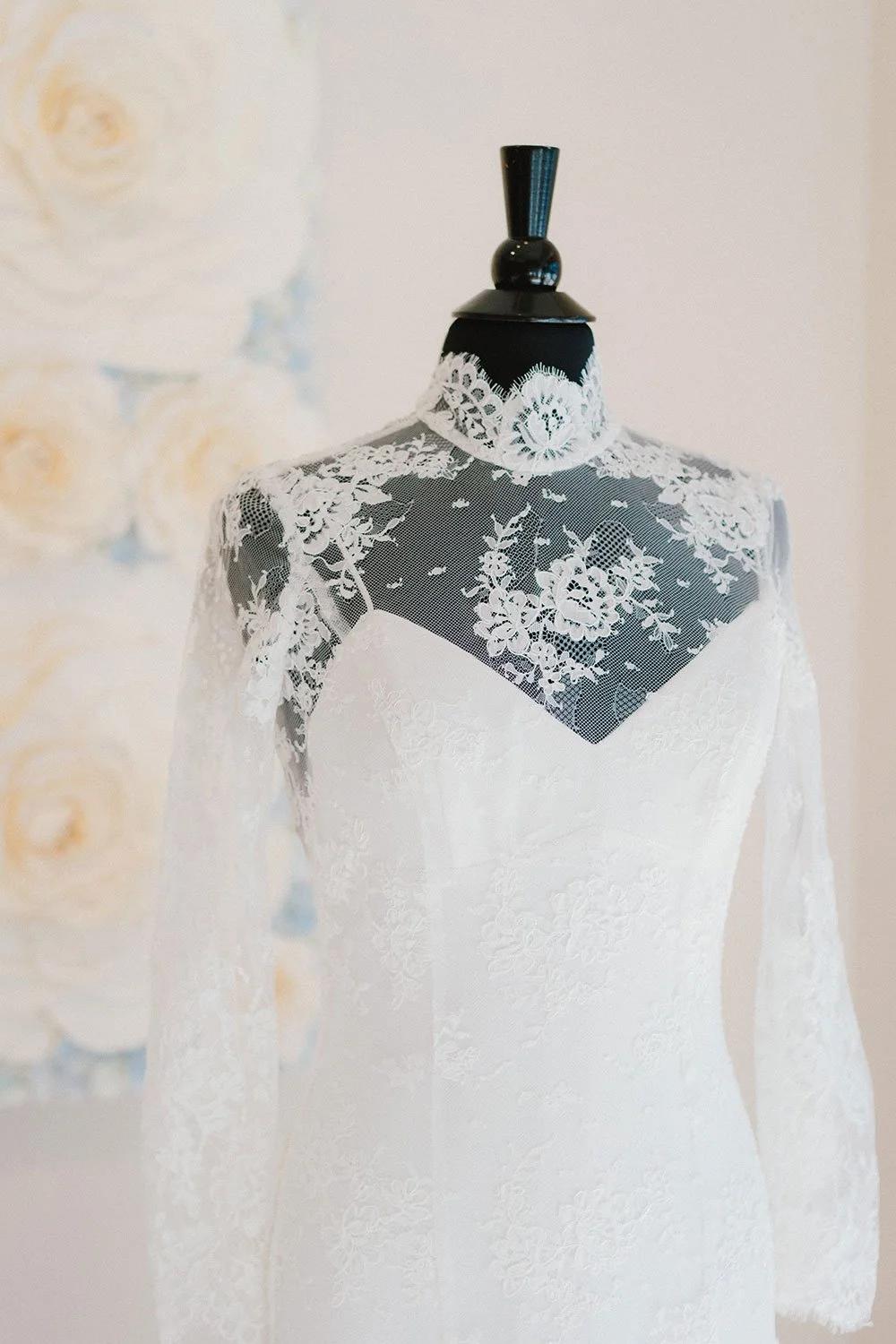 A mannequin dressed in a white lace wedding gown with floral lace details and a sheer lace overlay on the shoulders and neckline.