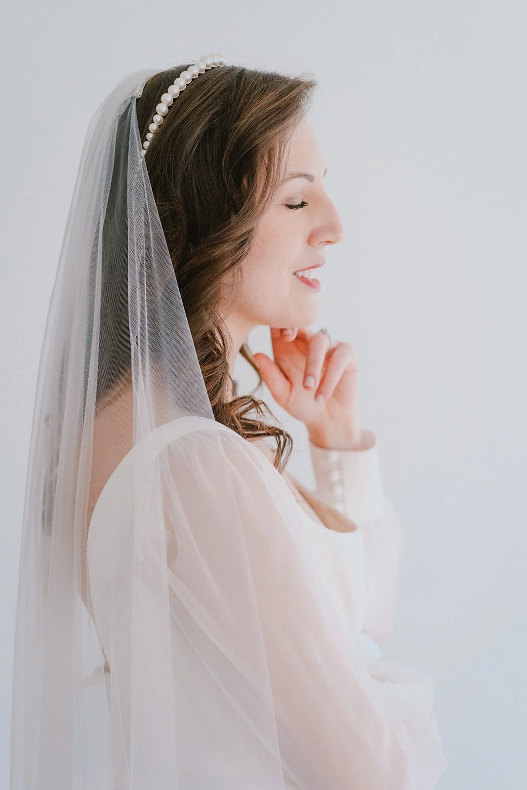 A bride with long dark hair wearing a pearl headband, a white veil, and a white dress, smiling softly with her eyes closed, facing right.