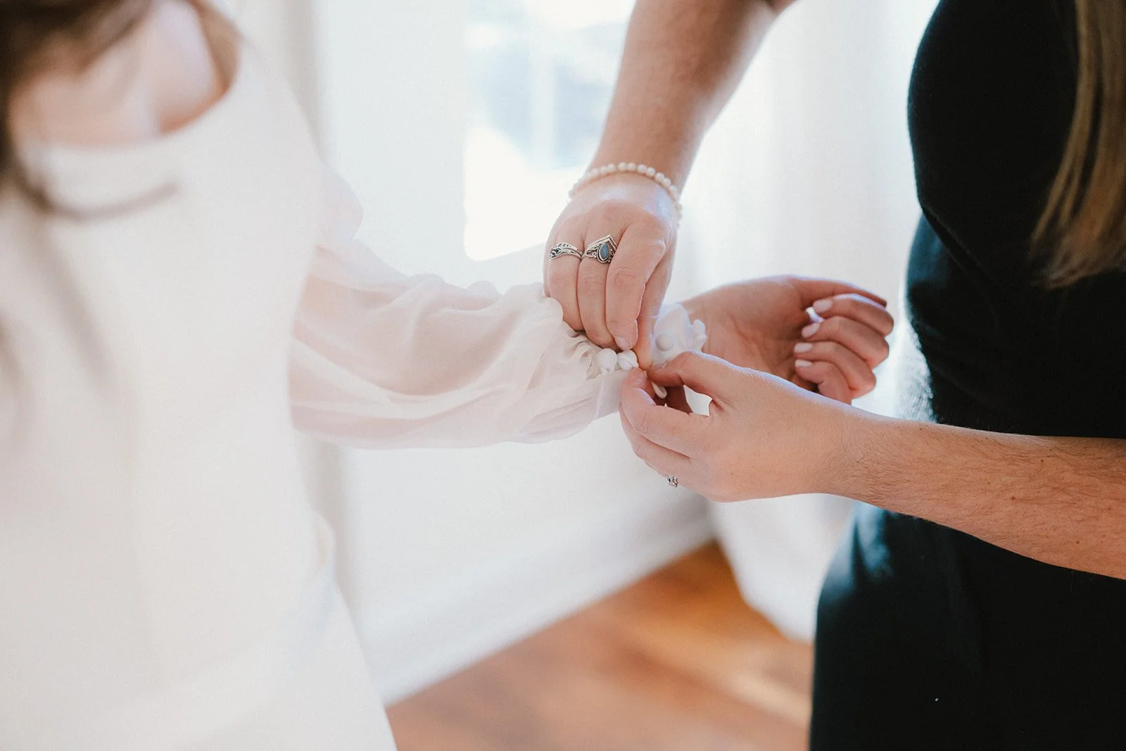 Two people holding a child's hand, helping them with buttons on a white dress, with a focus on their hands and jewelry.