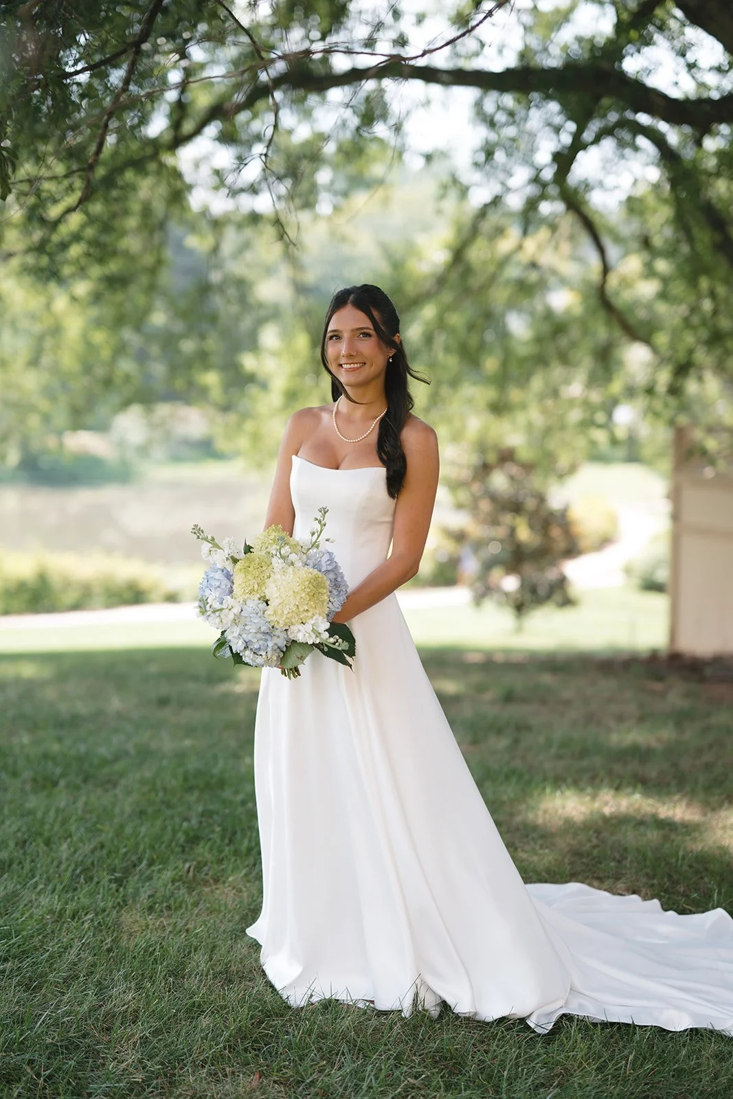 A bride in a white wedding dress holding a bouquet of flowers outdoors under a tree with green leaves.