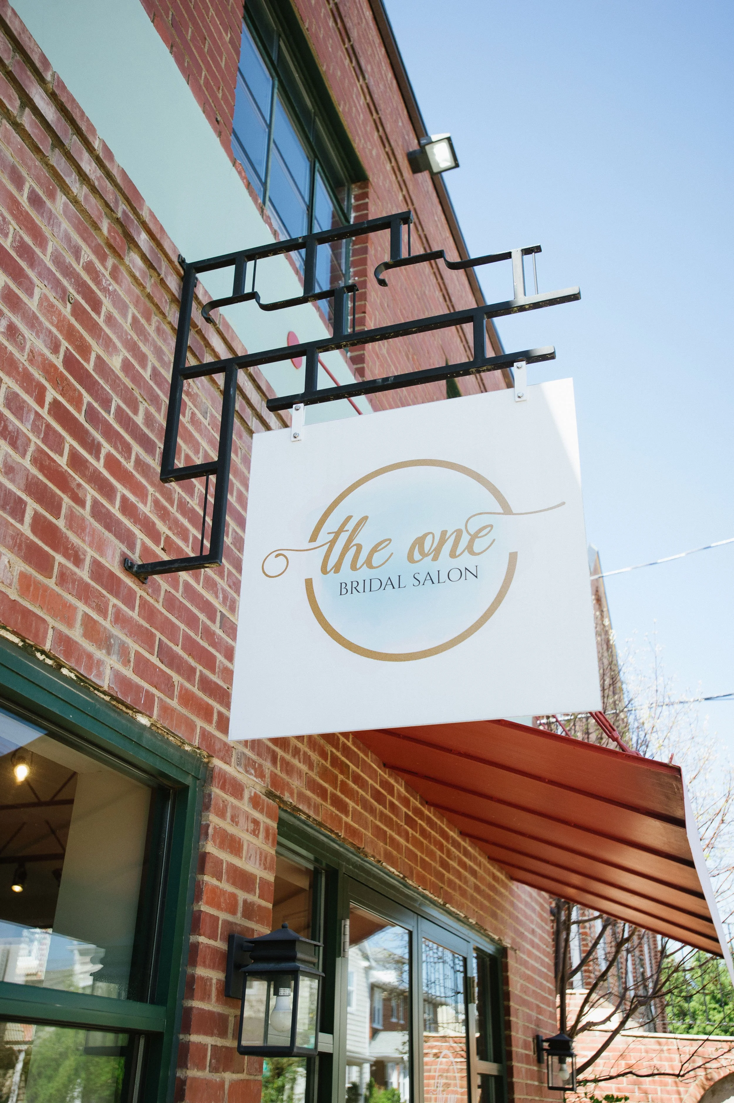Exterior view of a brick building with large windows, a sign that reads "the one BRIDAL SALON," black metal ornamental brackets, and outdoor wall lamps, under a red awning, with clear sky and trees in the background.