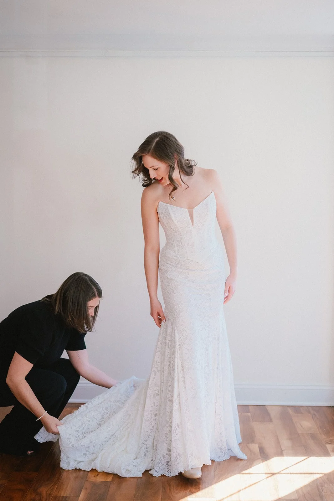 A woman in a white wedding dress is being assisted by another woman as she prepares for her wedding. The bride is looking down and smiling, with her dark hair styled in loose waves, standing on a wooden floor against a plain white wall.