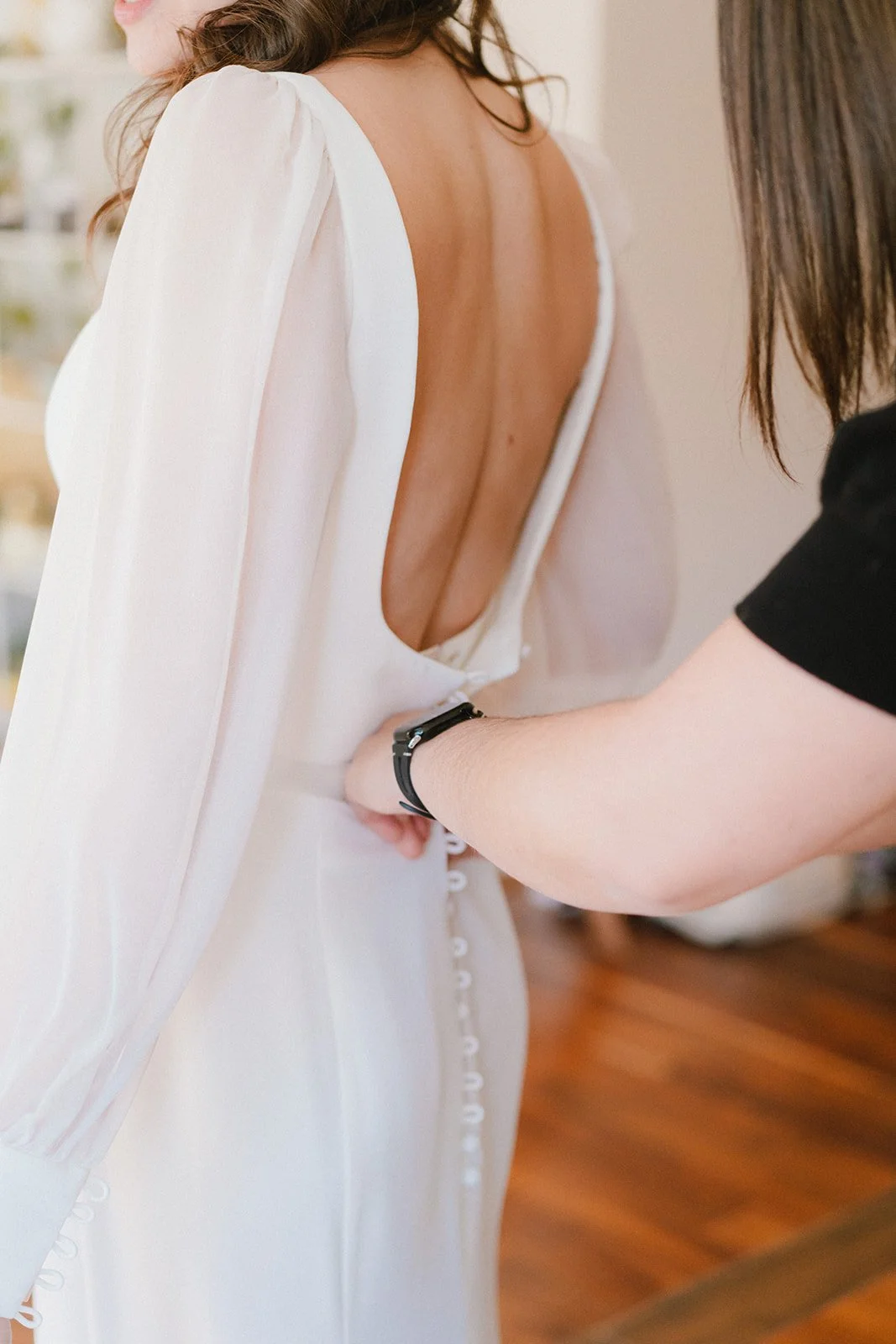 A woman is having her dress fastened by another person on her back, with the focus on the back of the dress with buttons and a deep open back design.