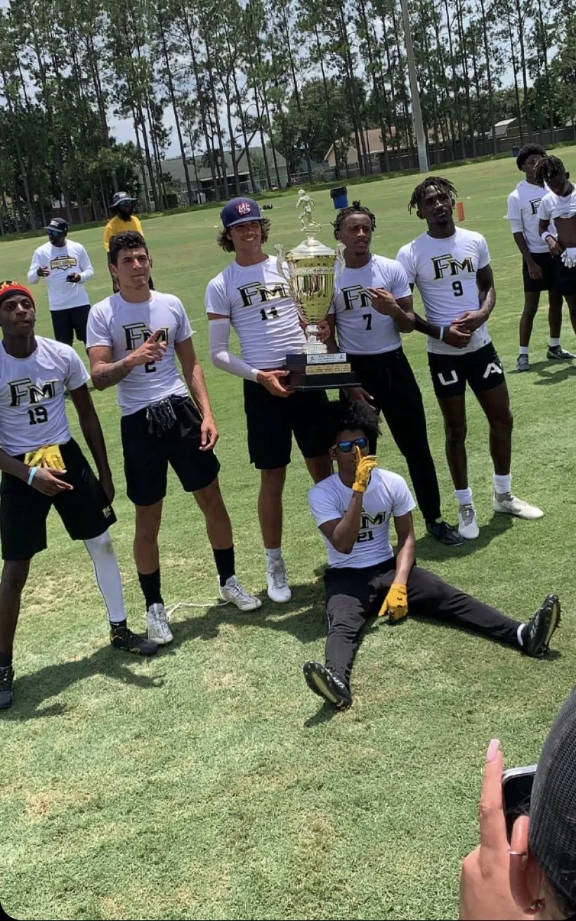 Group of young athletes, wearing sports jerseys with 'FM' logo, celebrating on a grassy field, holding a large trophy, with one person sitting on the ground making a gesture.