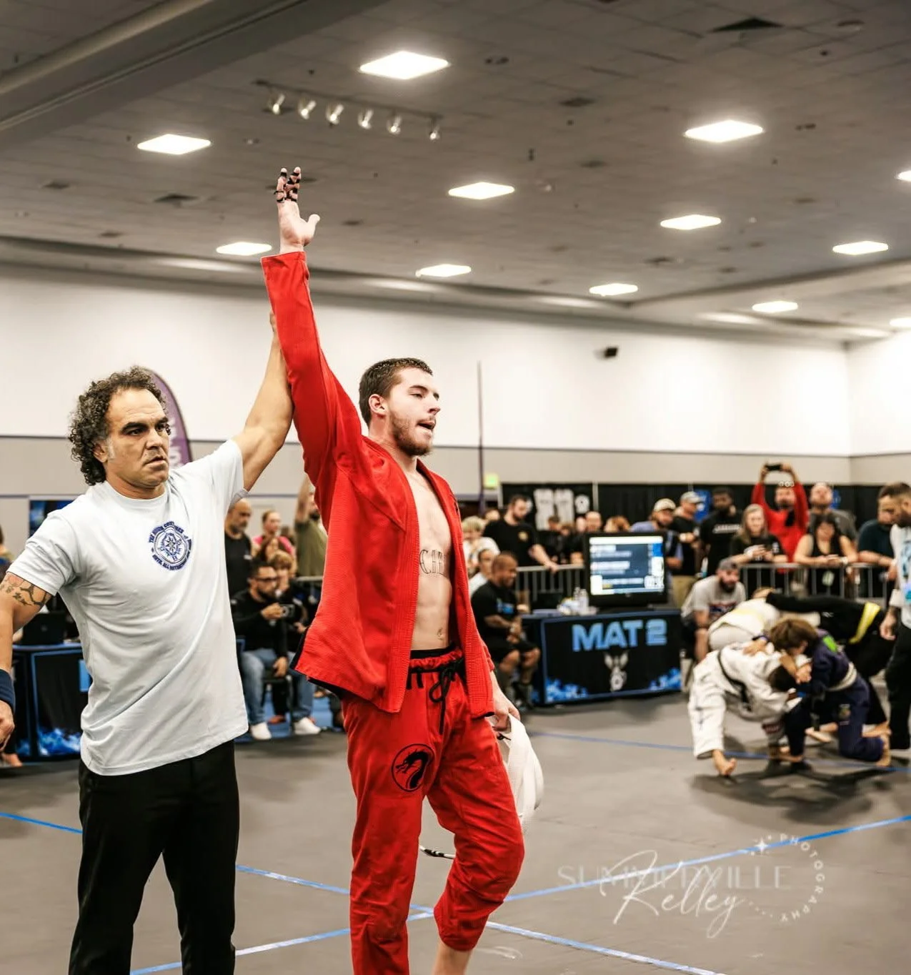 A male martial artist in red gi with a black dragon logo on pants, holding a white fight glove, stands with one arm raised as a referee, who has curly hair and tattoos, announces his victory in a martial arts competition surrounded by spectators, some in jujitsu gis, in a large indoor gym.
