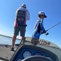 Two people are fishing from a boat on a lake, both wearing hats and casual clothing. One person is holding a fishing rod. The boat has a label that appears to read 'TATM...' and is equipped with electronic equipment.