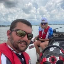 Two people on a boat, smiling, with one man in sunglasses in the foreground and a woman in the background wearing a hat and sunglasses, with water and cloudy sky behind them.