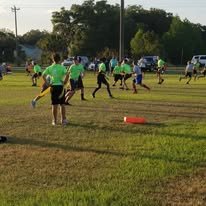 Children playing flag football on a grassy field during the day, with some people wearing green jerseys and others in blue and black.