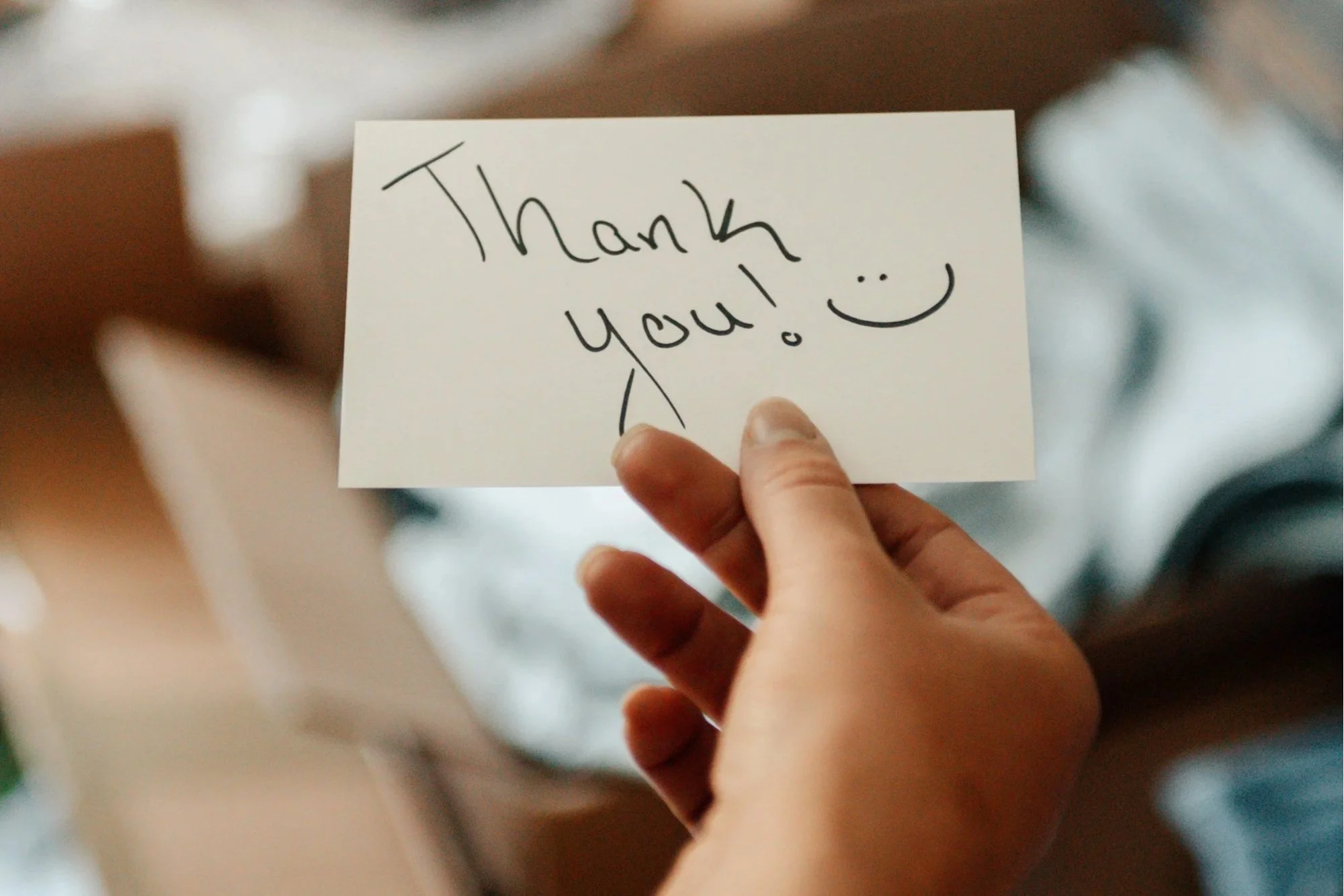Person holding a white card with a handwritten message 'Thank You!' and a smiley face, with a blurred background.