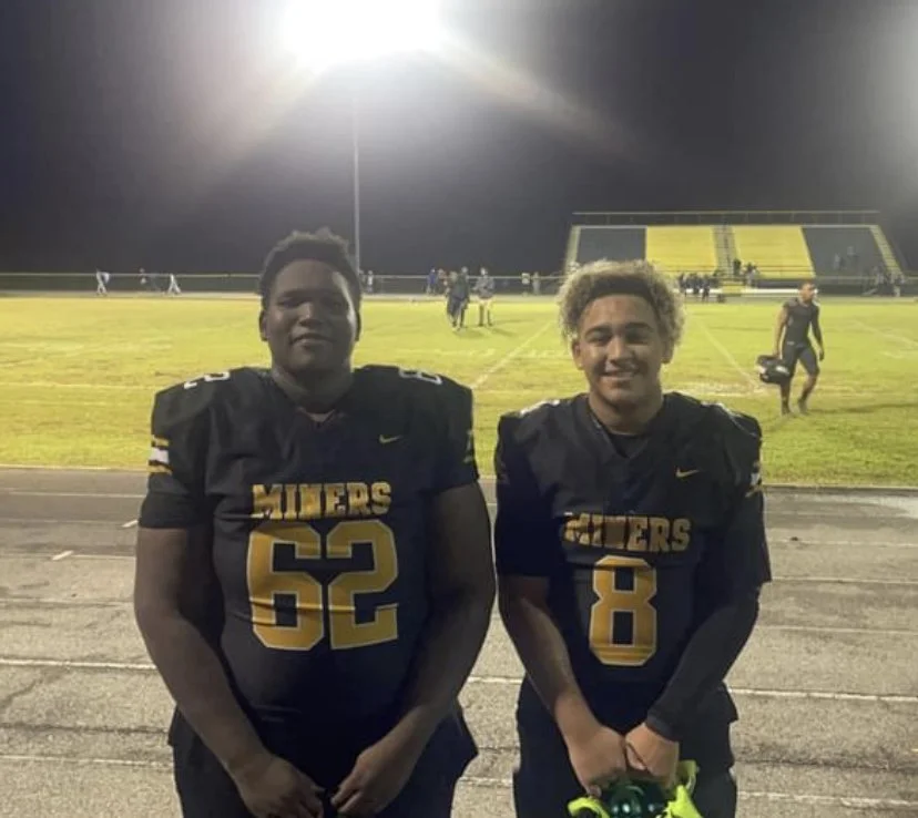 Two high school football players in black and yellow uniforms standing on a football field at night, smiling after a game.
