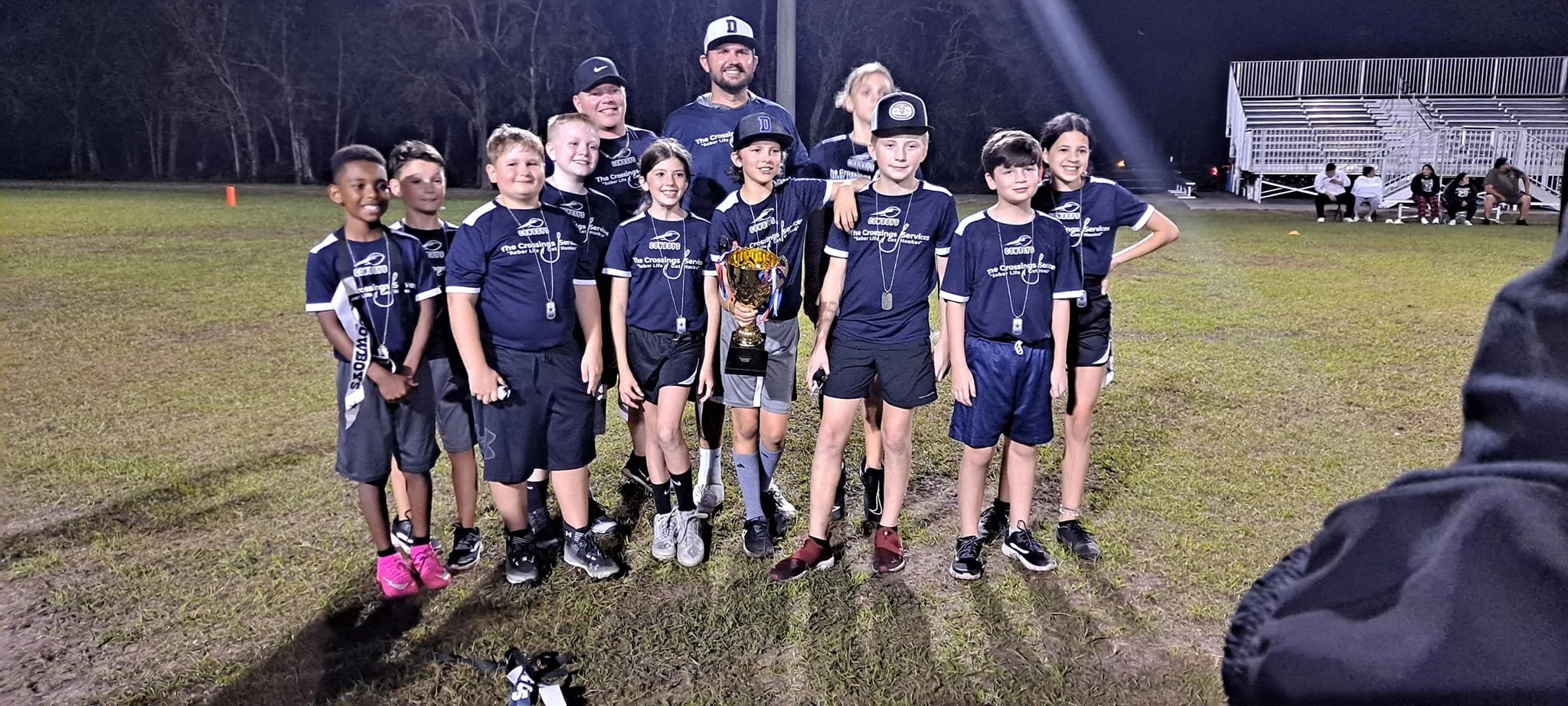 A group of young boys and girls in sports uniforms, along with two adult men, standing on a grassy field at night. They are celebrating with a trophy, some wearing medals, after a sports game.