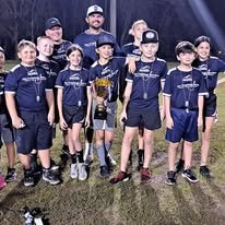 A group of young boys in baseball uniforms standing on a field, holding a trophy, with two adult coaches behind them.