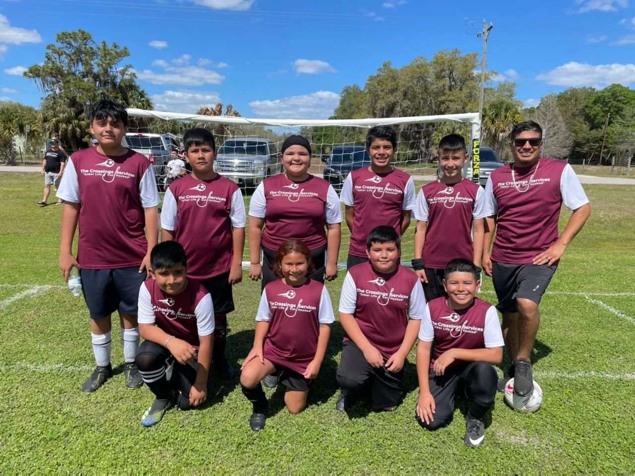 Youth soccer team in maroon and white uniforms posing in front of a soccer goal on a grassy field, with coaches and trees in the background.