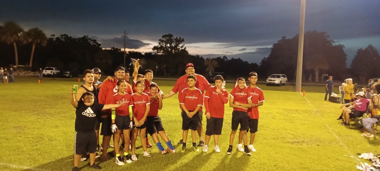 A youth sports team dressed in red jerseys and black shorts standing on a grassy field at dusk, with some team members speaking and others smiling, while people sit and stand along the side of the field in the background.