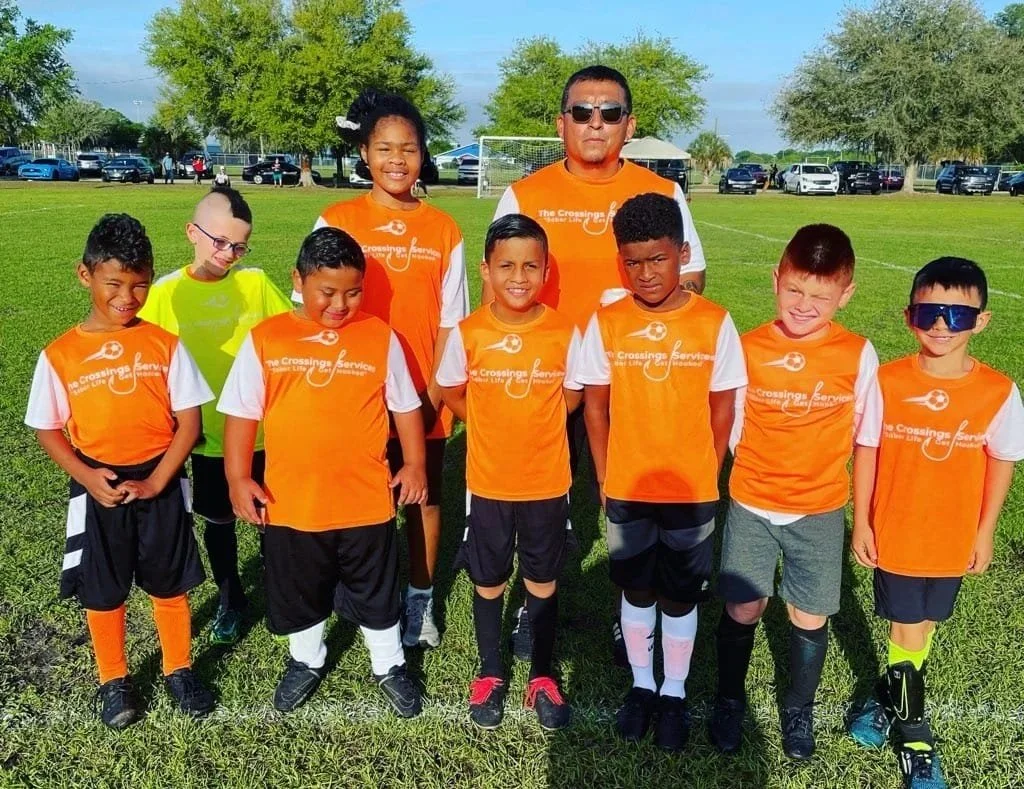 A youth soccer team with a coach in orange jerseys, standing on a grassy field with trees and parked cars in the background.