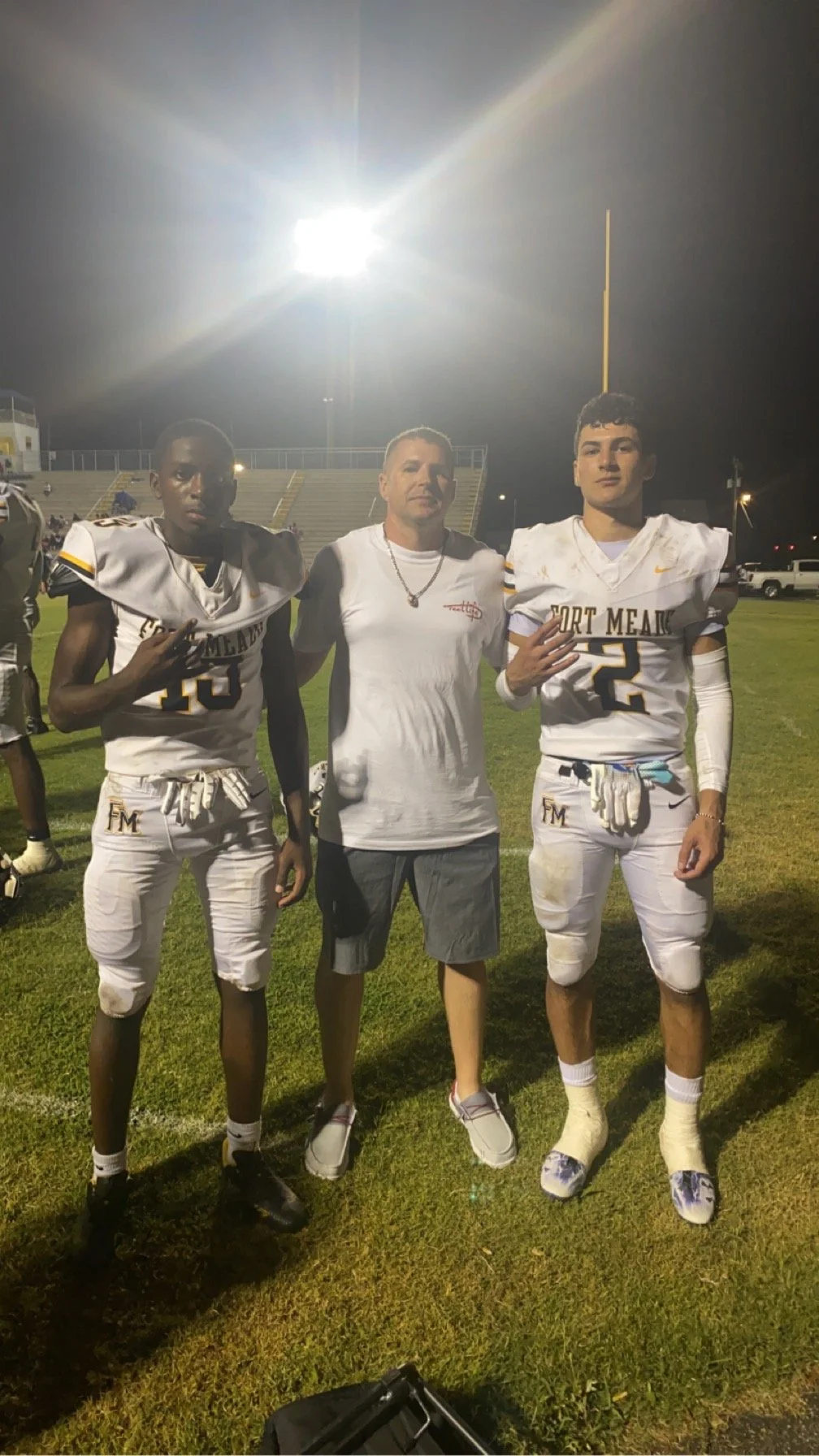 Two football players in white uniforms with black and yellow accents, posing with a coach or trainer in the middle, under stadium lights on a football field at night.
