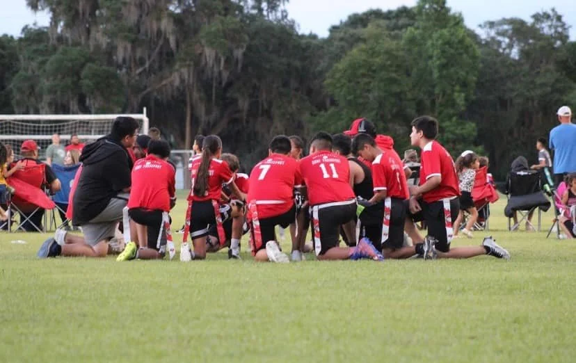 Youth football team kneeling on the field in a huddle during a game, surrounded by spectators and coaches.