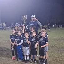 Group of young boys in soccer uniforms on a field, with a coach holding a trophy in the background at night.