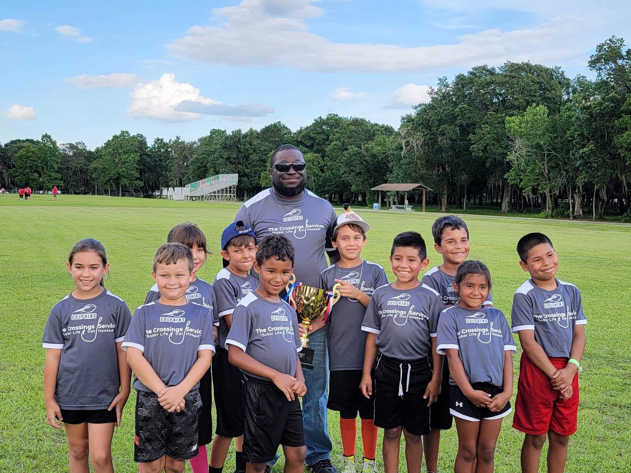 A group of children standing on a grassy field with a coach, holding a trophy, with trees and a blue sky with clouds in the background.