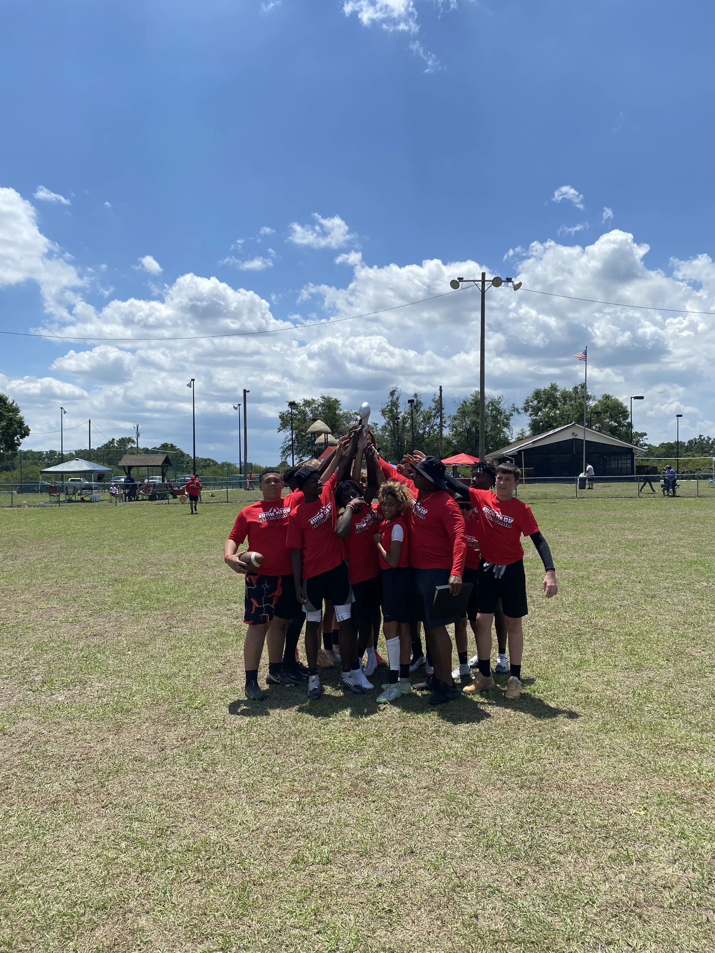 A group of young athletes in red shirts are standing outdoors on a sports field, holding a trophy together. The sky is mostly sunny with some clouds, and there are trees, a few structures, and people in the background.