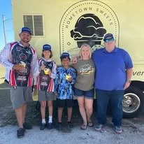 Group of five people, including two children, standing outside a building with a sign that reads "Fultown Sweet". They are smiling, and some are wearing baseball caps and jackets.