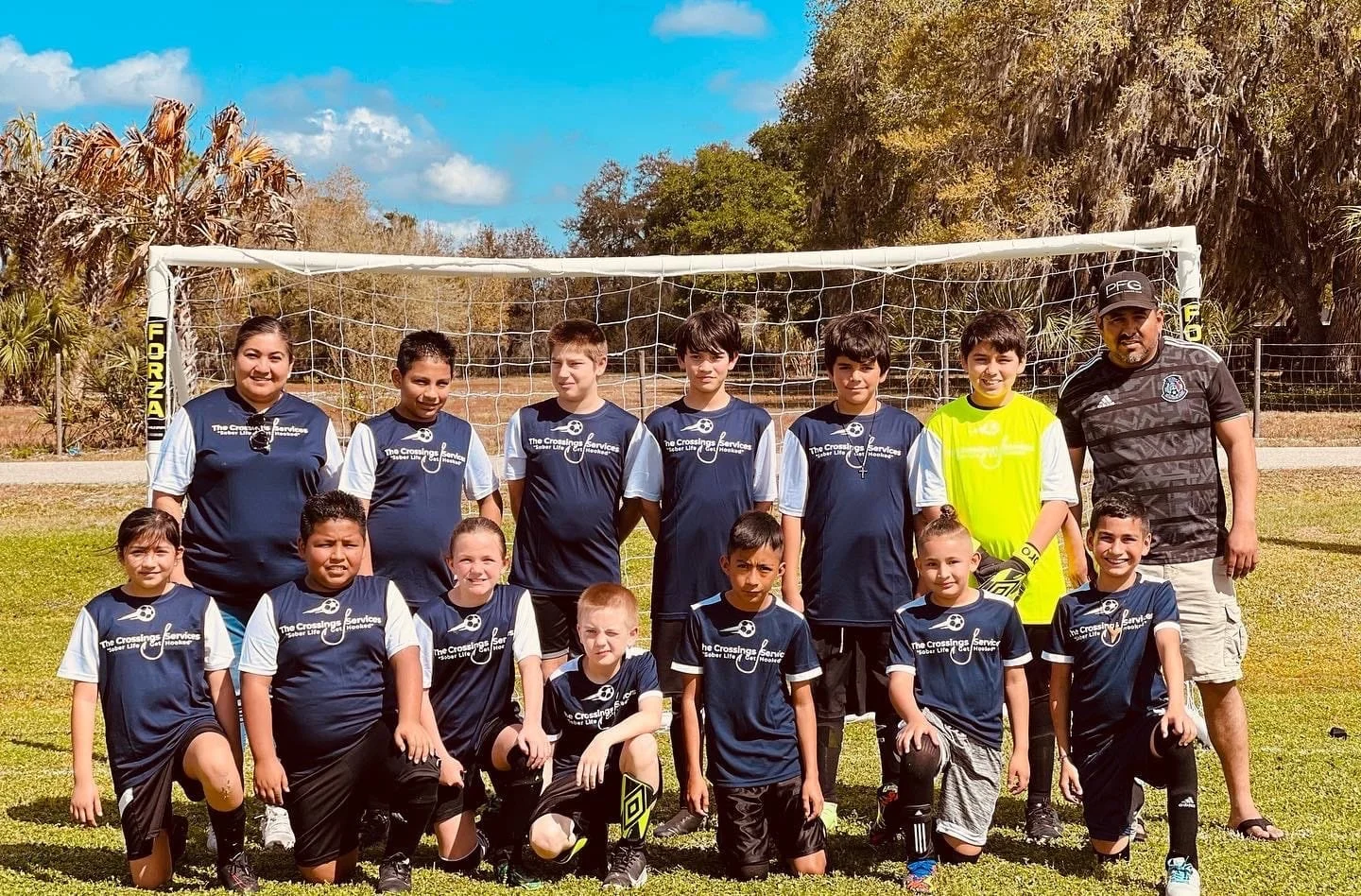 Group of young children and adults posing on a soccer field in front of a soccer goal. The children are wearing navy blue and white soccer uniforms, and the adults are dressed casually, with one wearing a black sports shirt and the other a navy blue shirt. The scene is outdoors on a sunny day with trees and a blue sky in the background.