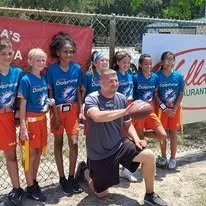 A man kneeling in front of a group of young girls in sports uniforms holding footballs, on a baseball field with a chain-link fence in the background.