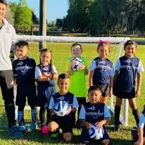 Group of six young children in soccer uniforms posing on a field, with one adult standing nearby.