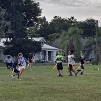 Group of children playing soccer outdoors on a grassy field with trees and a building in the background.