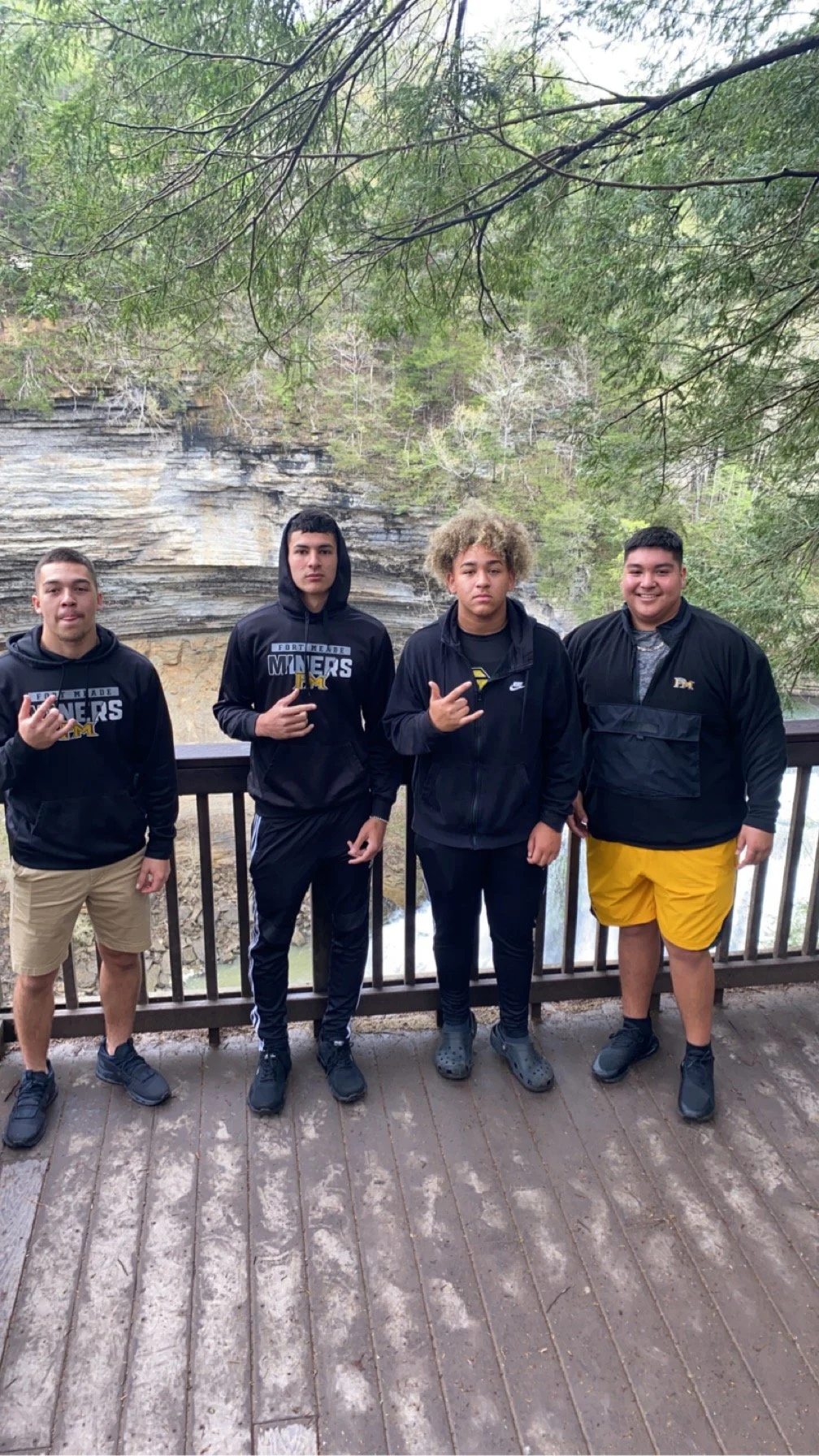 Four teenagers standing on a wooden platform in front of a railing with a river and rocky cliff in the background, surrounded by trees.
