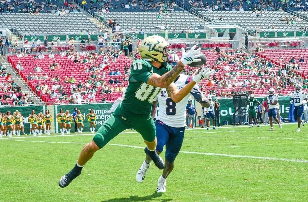 Football player in green uniform catching a football in a stadium with spectators and cheerleaders in the background.