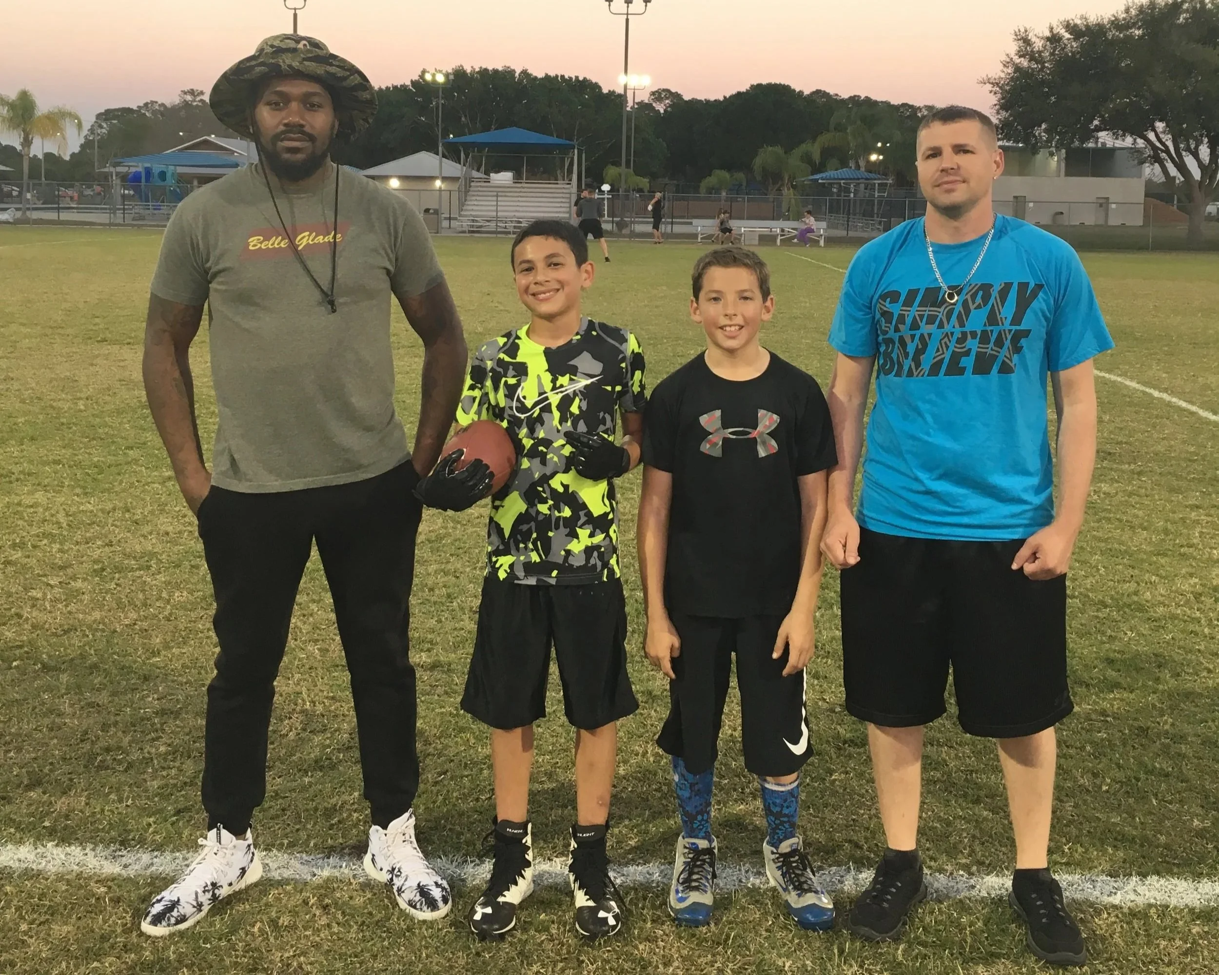 Four males and two young boys standing on a football field at sunset, with a playground, trees, and a sports field in the background.