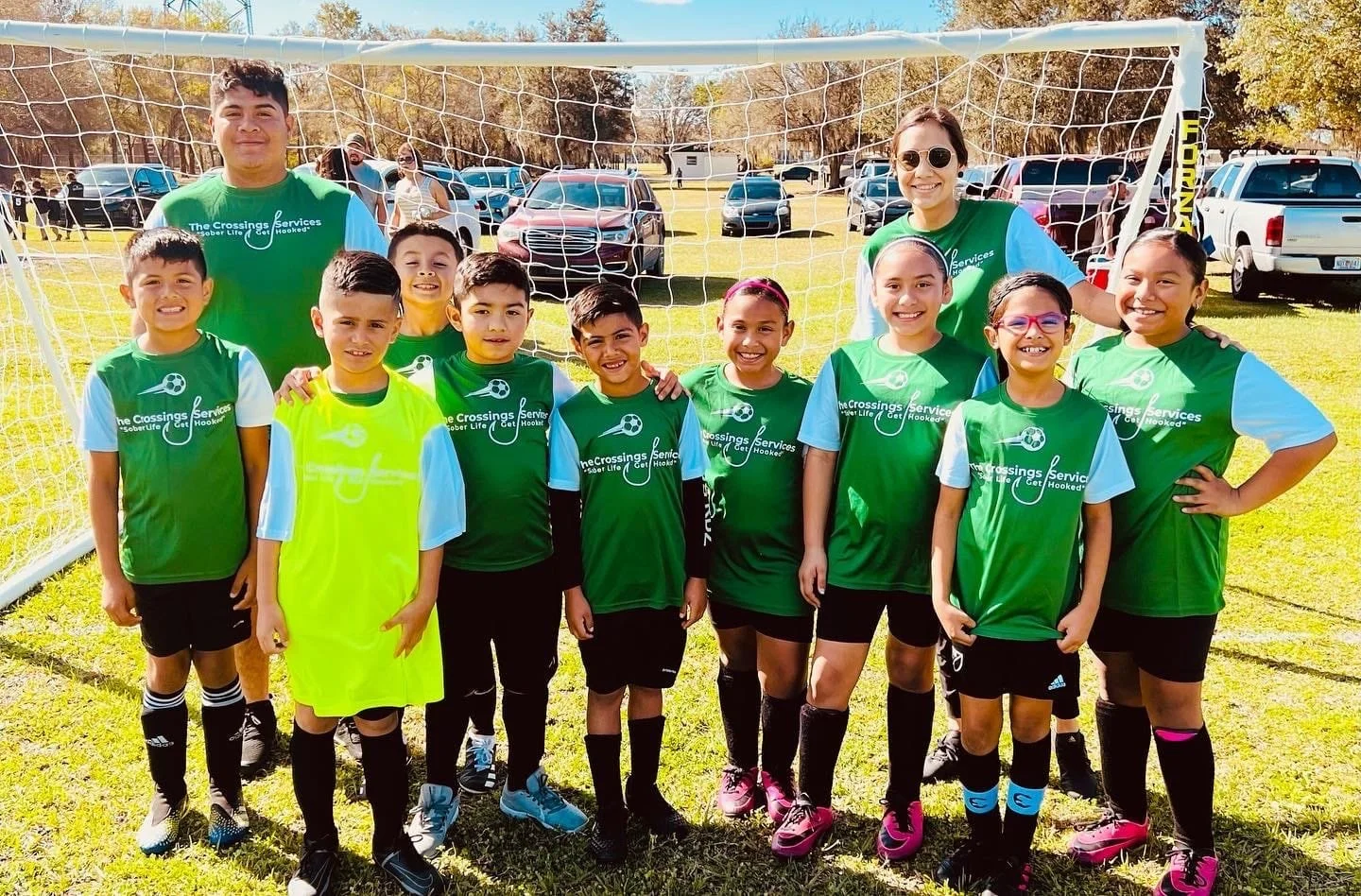 A group of young soccer players and their coaches standing in front of a soccer goal on a sunny day, smiling at the camera.