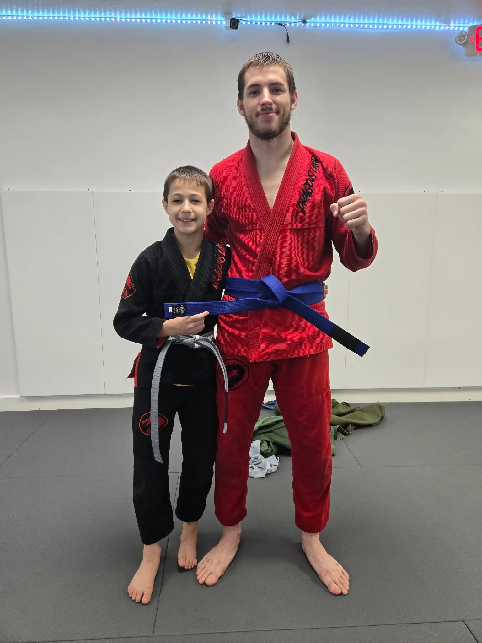 Two males wearing martial arts uniforms, standing together on a mat in a dojo. The taller man is in a red gi with a blue belt, and the shorter man is in a black gi with a gray belt. They are both barefoot, smiling, and posing together.