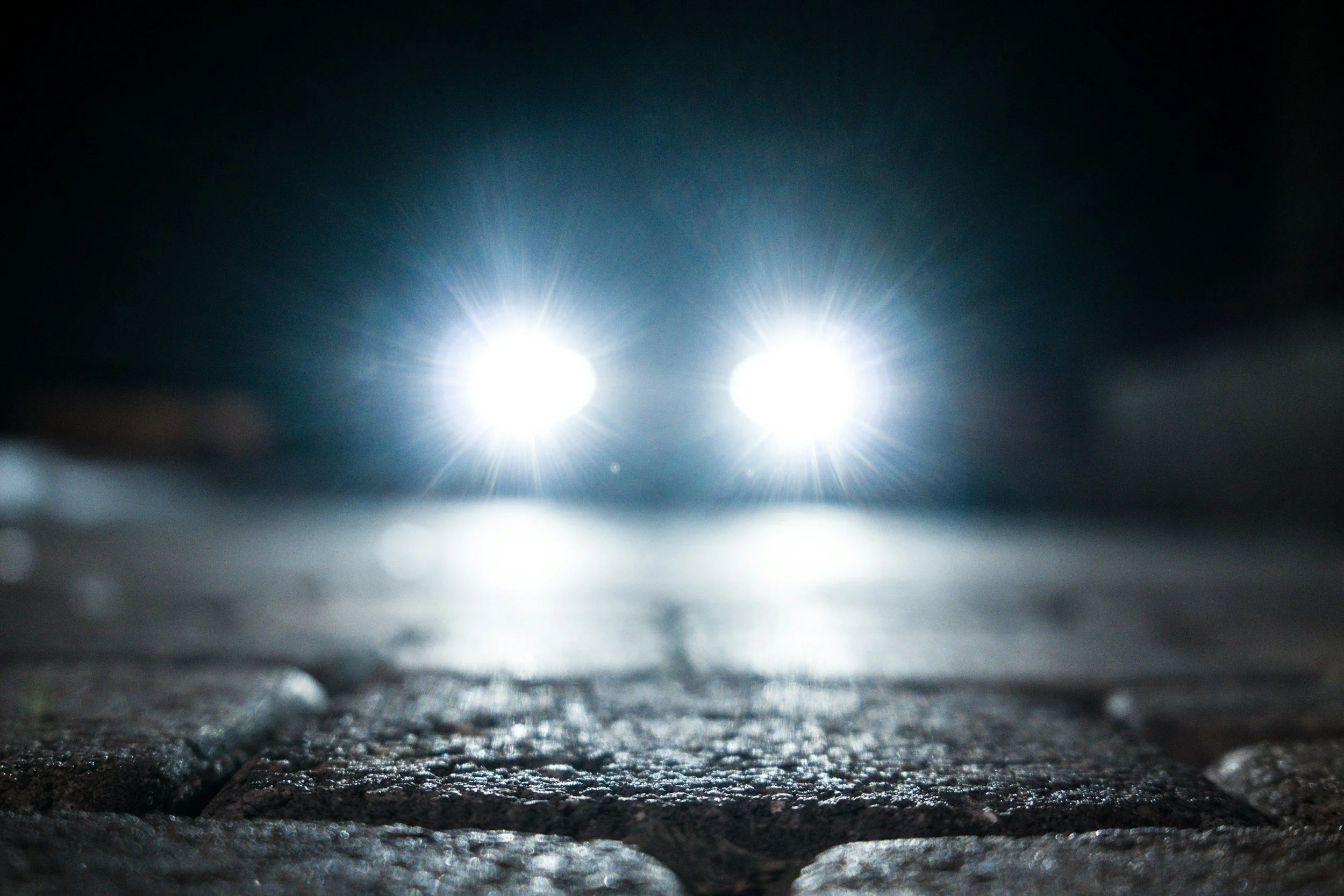Close-up of two bright car headlights shining on a textured, wet road at night.