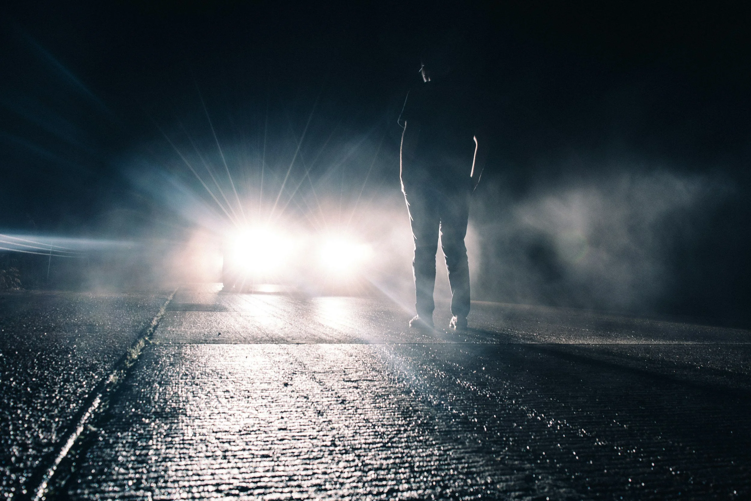 Person walking on a wet road at night with bright lights behind them, creating a silhouette and flare.