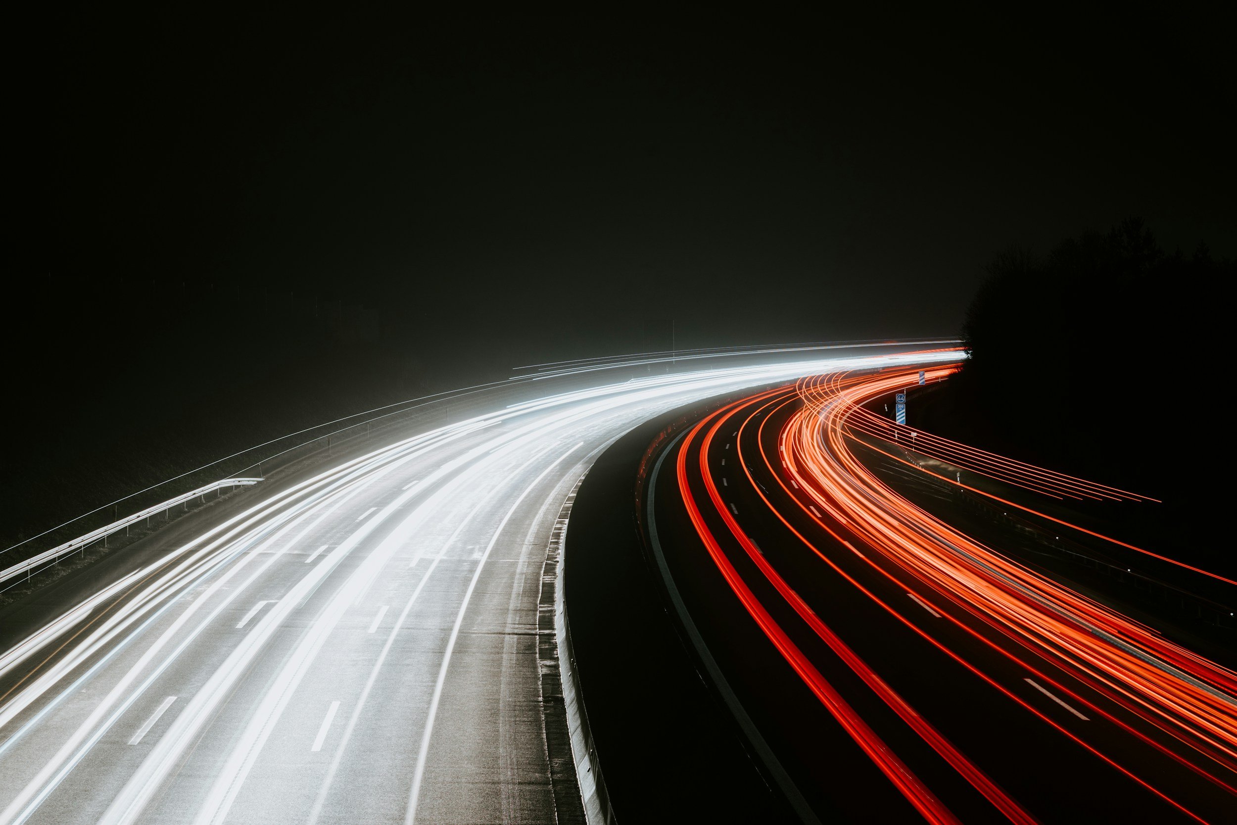 Nighttime photo of a highway with light trails from moving vehicles, showing white lights on one side and red lights on the other side, curving to the right.