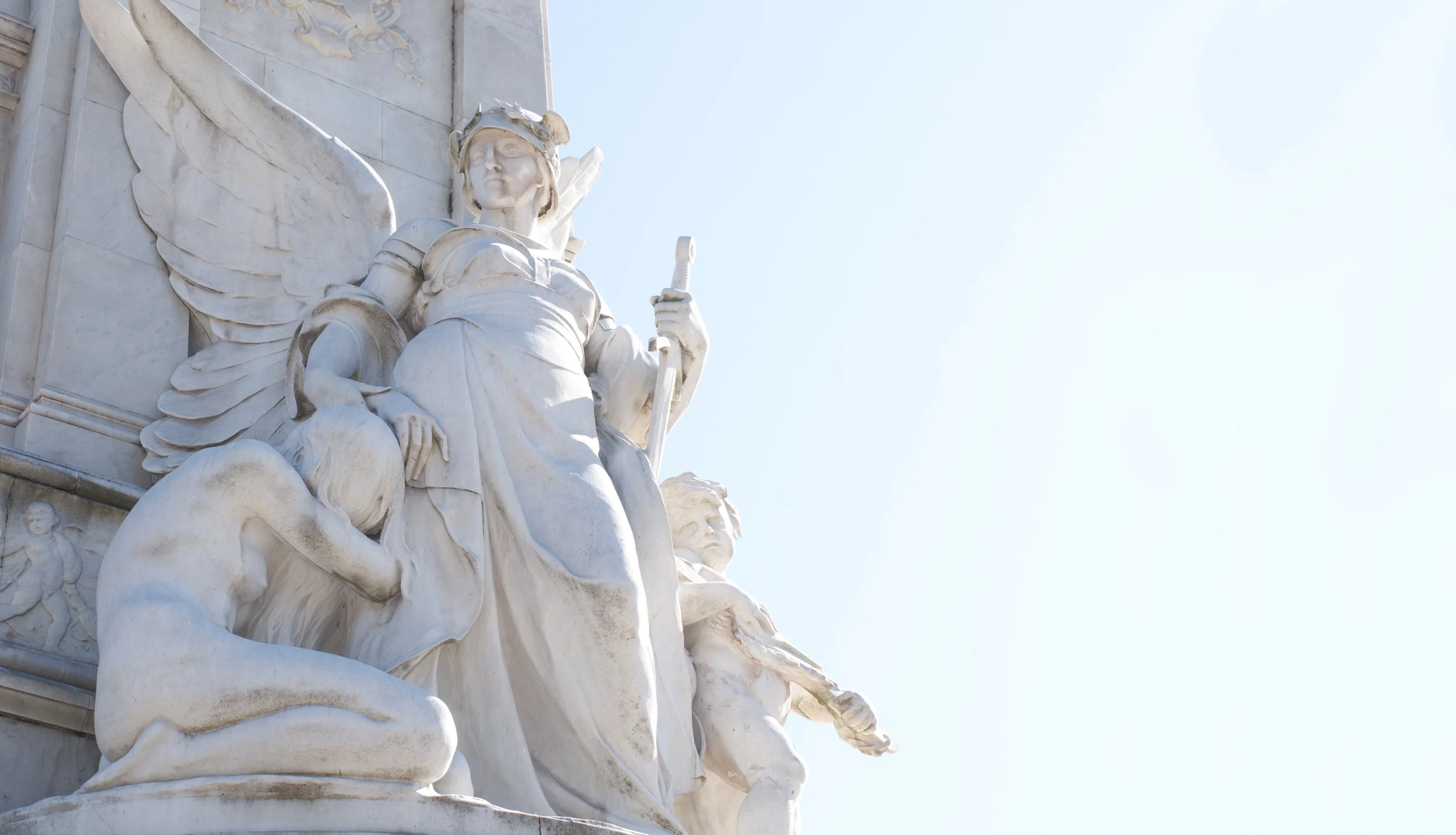 A close-up view of a white marble statue of an angel holding a sword, with detailed wings, part of a larger monument against a clear blue sky.