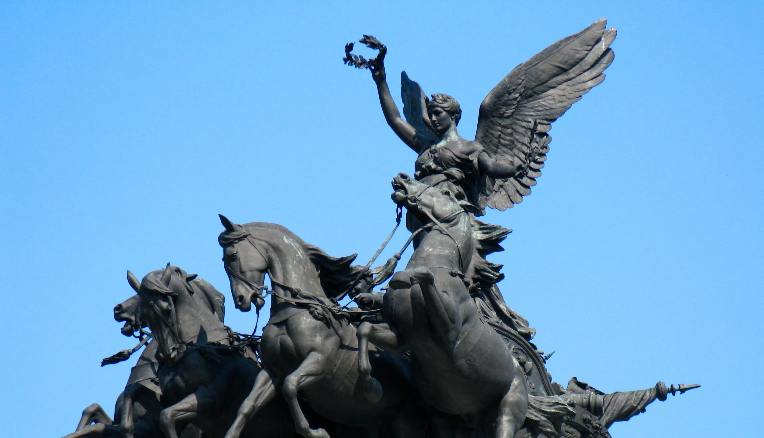A bronze statue of a woman riding a chariot pulled by horses, holding a laurel wreath in her raised hand, set against a blue sky.