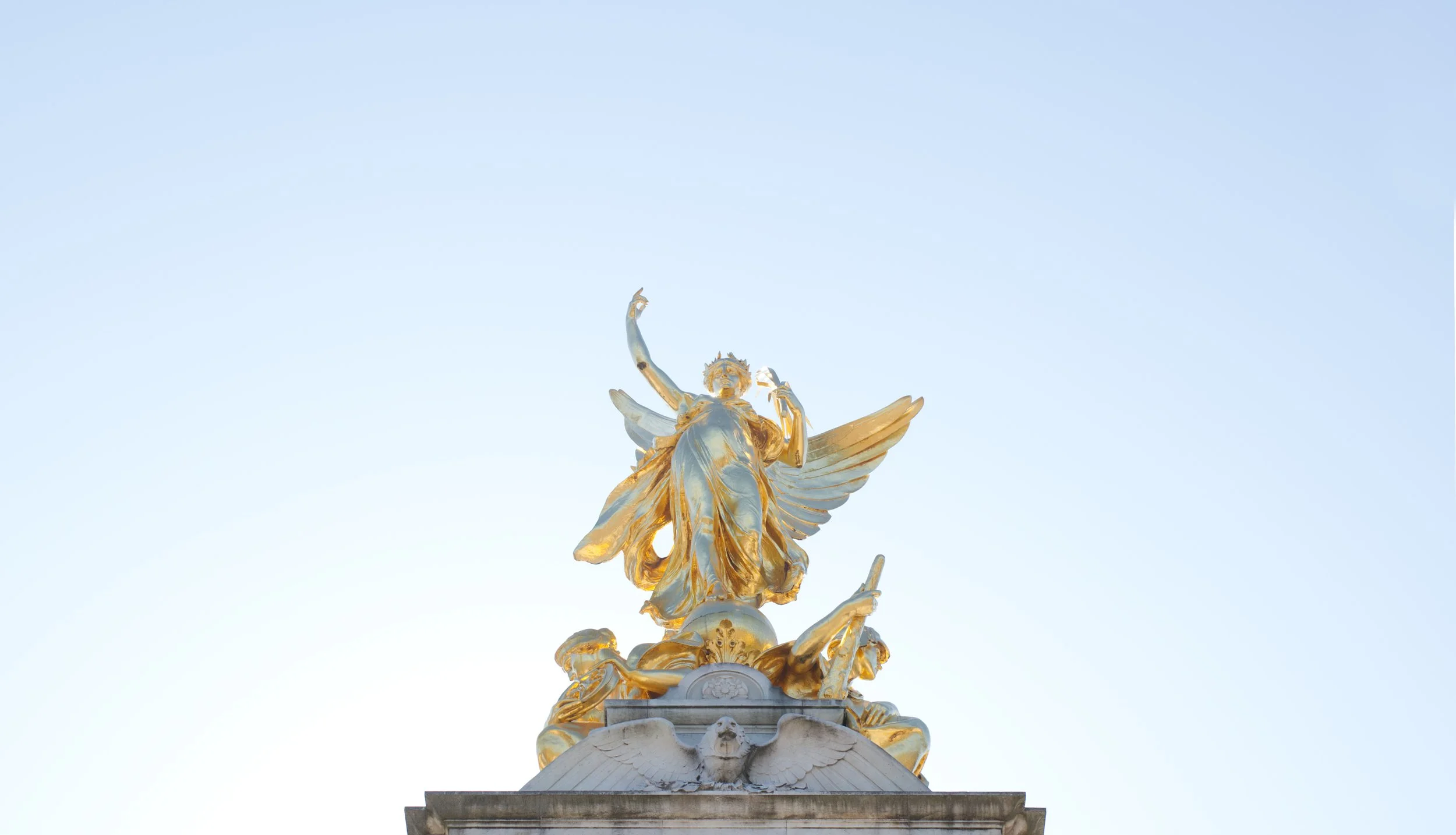 A golden statue of a winged figure holding a sword, standing atop a stone pedestal against a clear sky.