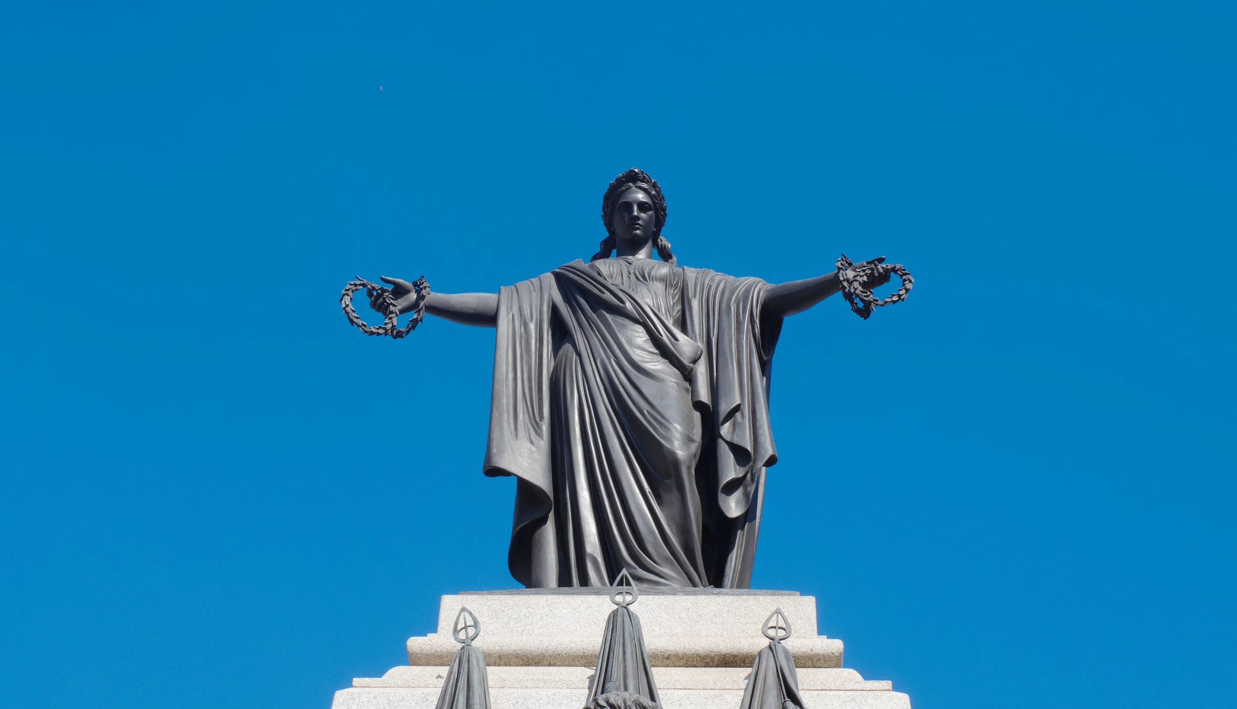 Statue of Lady Liberty with arms outstretched, holding a laurel wreath in each hand, against a clear blue sky.