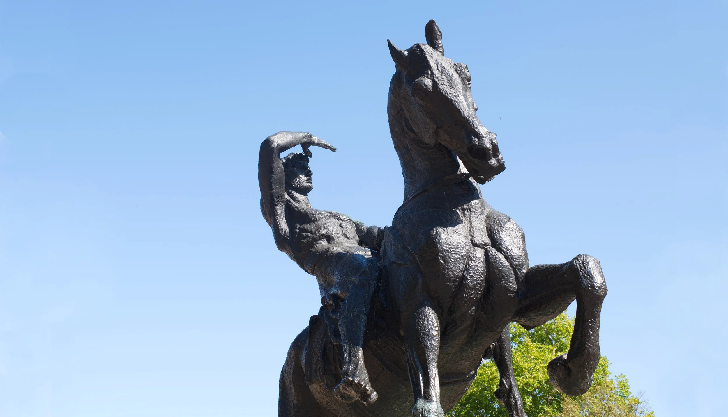 Bronze statue of a Native American man riding a horse against a clear blue sky, with green trees in the background.
